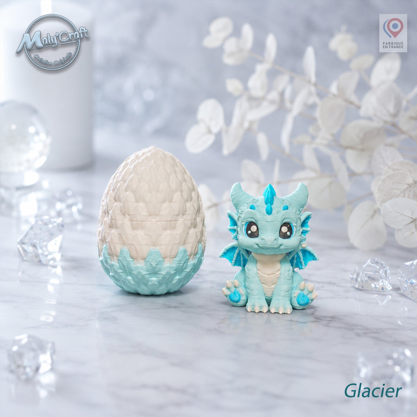 Figurine bébé dragon Glacier bleu glacier et blanc avec son œuf bicolore blanc et bleu, posés sur un marbre blanc. Création originale MalyCraft3D, impression 3D de précision réalisée en France. Dragon kawaii multicolore avec détails écailles. Ambiance glacée et féerique avec cristaux transparents, bougie blanche et branches d'eucalyptus séchées en arrière-plan.