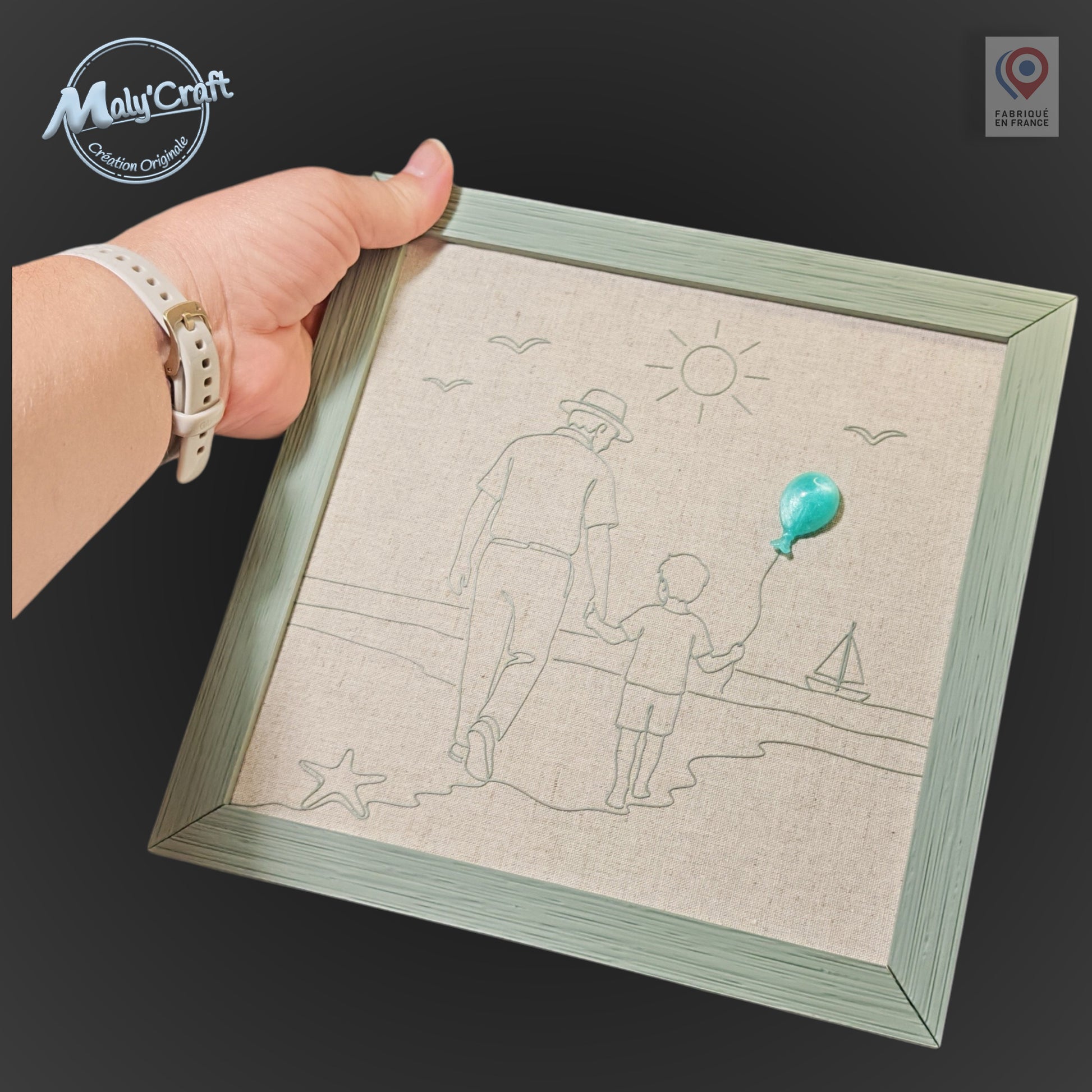 Peut inclure: Un cadre mural carré avec une bordure en bois vert clair et un fond en tissu beige. L'œuvre présente un dessin au trait représentant un adulte et un enfant marchant sur une plage, agrémenté d'un petit ballon turquoise en relief fixé dessus.