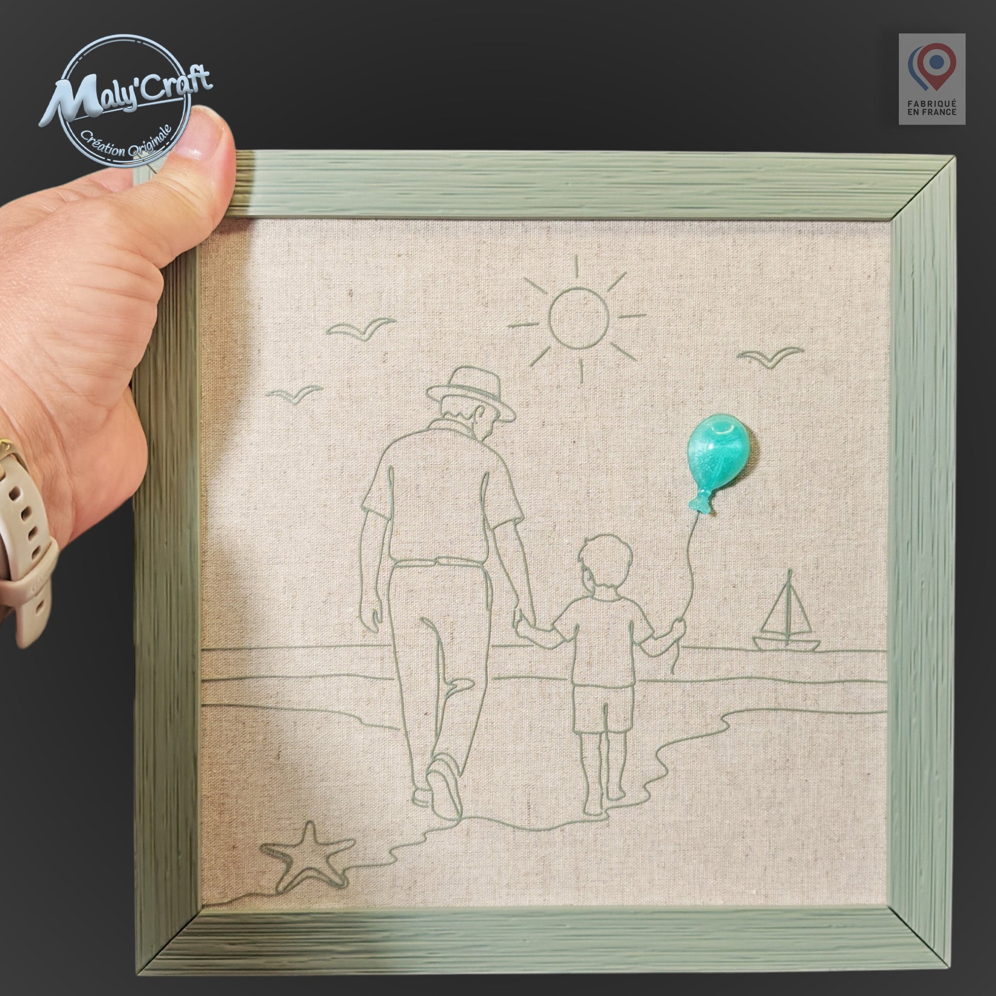 Peut inclure: Une décoration murale carrée encadrée présentant un dessin au trait noir sur un fond en tissu beige clair. L'illustration représente un adulte et un enfant marchant sur une plage, avec un petit ballon turquoise en relief sur la main.