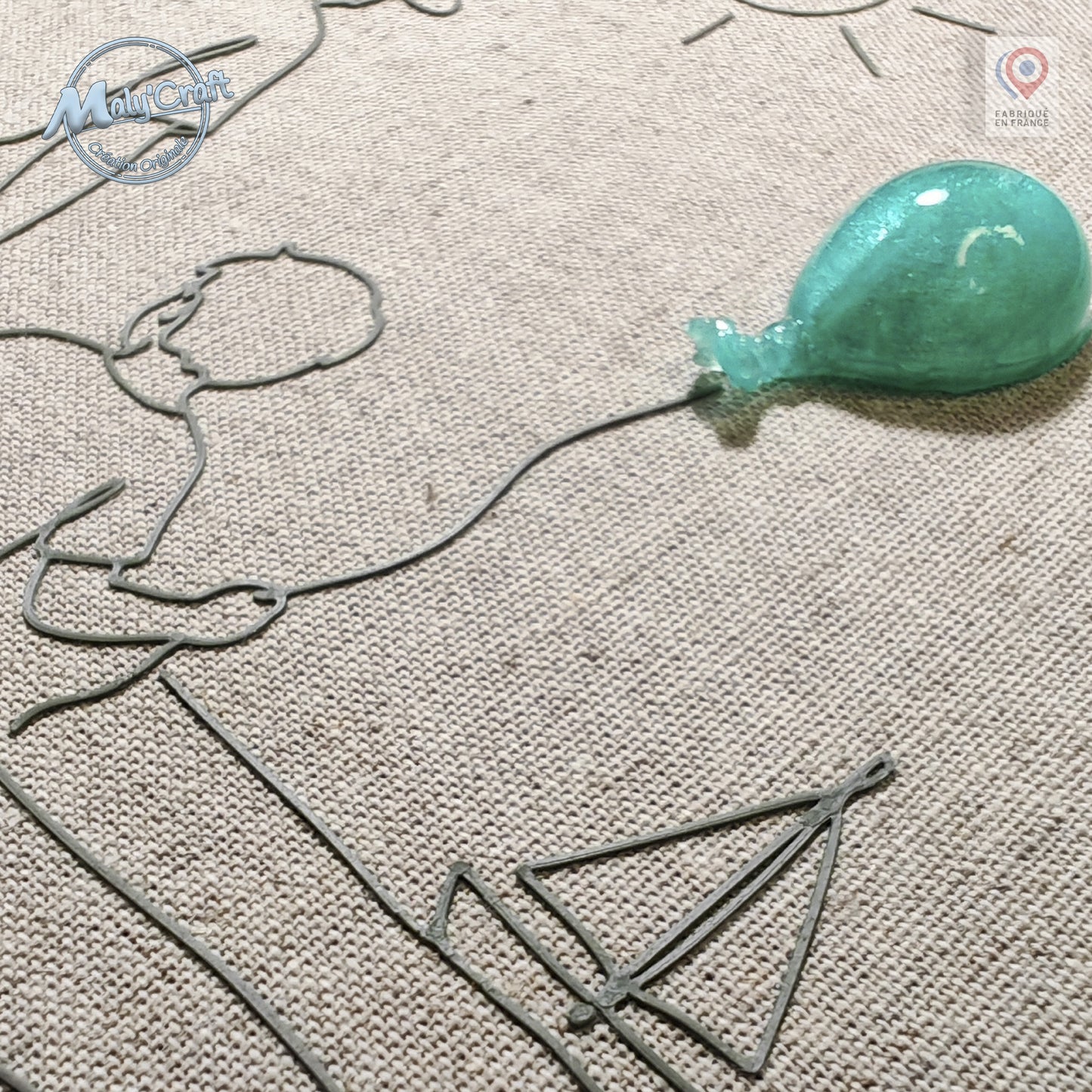 Peut inclure: Une décoration murale composée d'un contour en fil métallique représentant une personne tenant une ficelle reliée à un ballon bleu sarcelle translucide et brillant. L'œuvre est fixée sur un tissu beige texturé avec un voilier en fil.