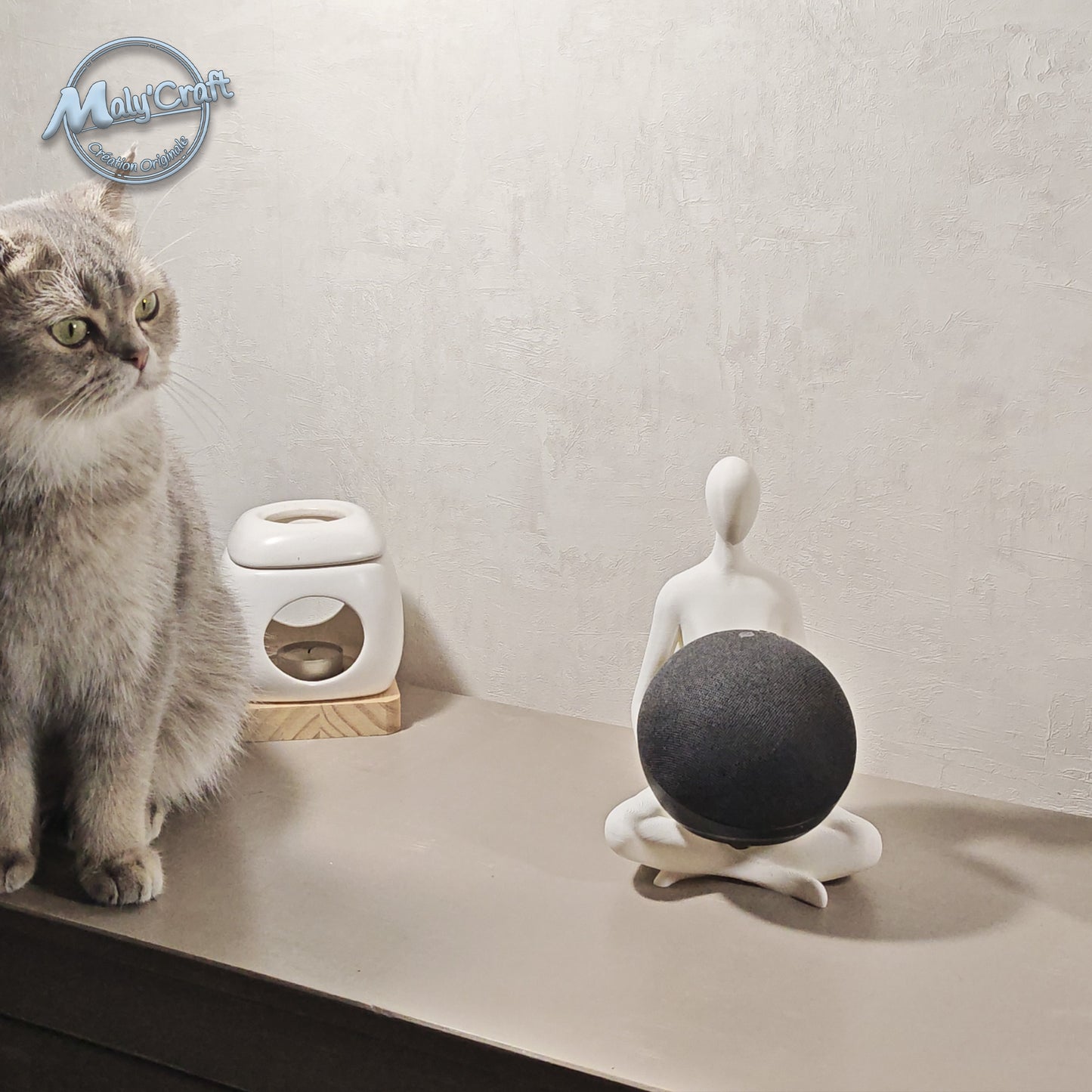 Support Alexa Echo Dot imprimé en 3D en forme de figurine lotus, posé sur un meuble aux côtés d’un chat gris. Design minimaliste et zen, fabriqué en France.
3D printed Echo Dot stand shaped as a lotus pose figurine, placed on a shelf next to a grey cat. Minimalist and zen design, handmade in France.