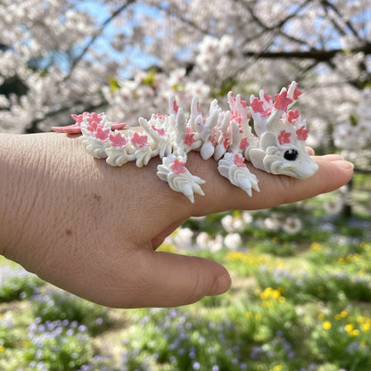 Dragon articulé Sakura blanc et rose, création Cinderwing3D, imprimé en 3D par Maly’Craft. Figurine flexible inspirée des fleurs de cerisier, présentée dans différents décors naturels et sur fond uni. Idéale pour la décoration, la collection ou un cadeau poétique aux tons printaniers.