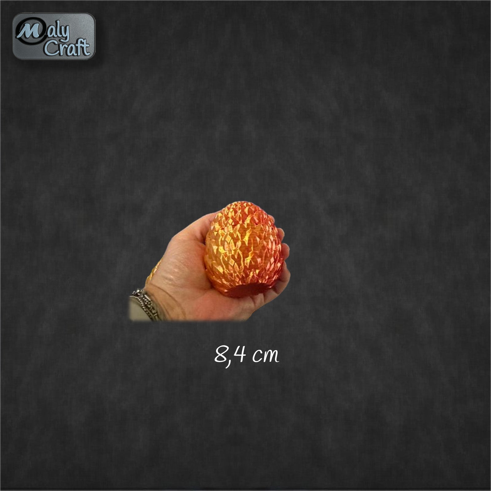 Œuf de dragon orange et doré imprimé en 3D, tenu dans la main pour montrer sa taille (8,4 cm). Création Cinderwing3D imprimée par Maly’Craft, au rendu irisé et détaillé.