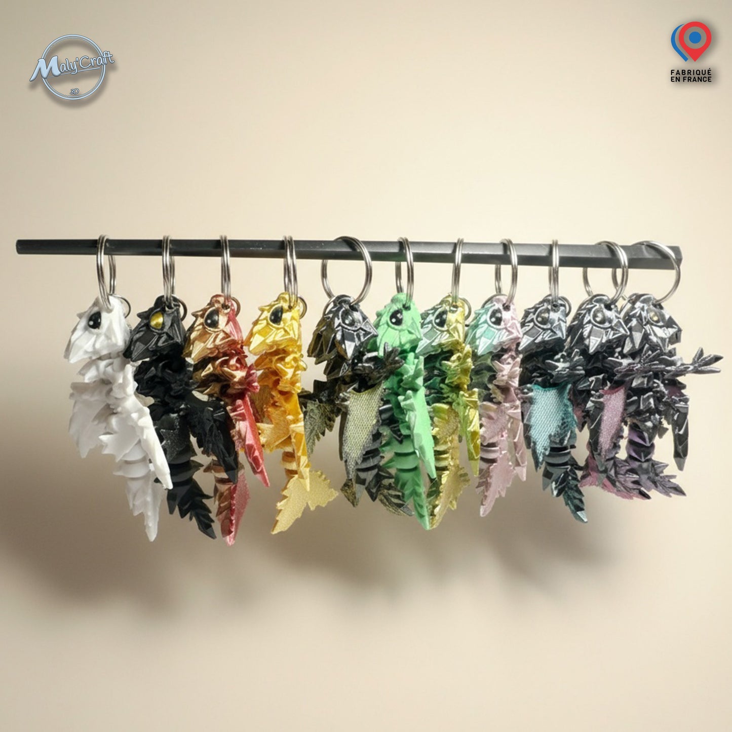 Ensemble de porte-clés dragon articulés imprimés en 3D, présentés côte à côte en plusieurs couleurs brillantes (blanc, noir, or, vert, rose, etc.). Chaque dragon au design détaillé évoque l’univers fantasy et médiéval. Idéal pour les fans de dragons, de jeux de rôle, ou d’univers comme Donjons & Dragons et Game of Thrones. Parfait comme cadeau geek, original et artisanal. Logo MalyCraft visible en haut à gauche.