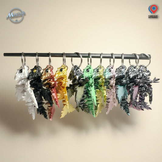 Ensemble de porte-clés dragon articulés imprimés en 3D, présentés côte à côte en plusieurs couleurs brillantes (blanc, noir, or, vert, rose, etc.). Chaque dragon au design détaillé évoque l’univers fantasy et médiéval. Idéal pour les fans de dragons, de jeux de rôle, ou d’univers comme Donjons & Dragons et Game of Thrones. Parfait comme cadeau geek, original et artisanal. Logo MalyCraft visible en haut à gauche.