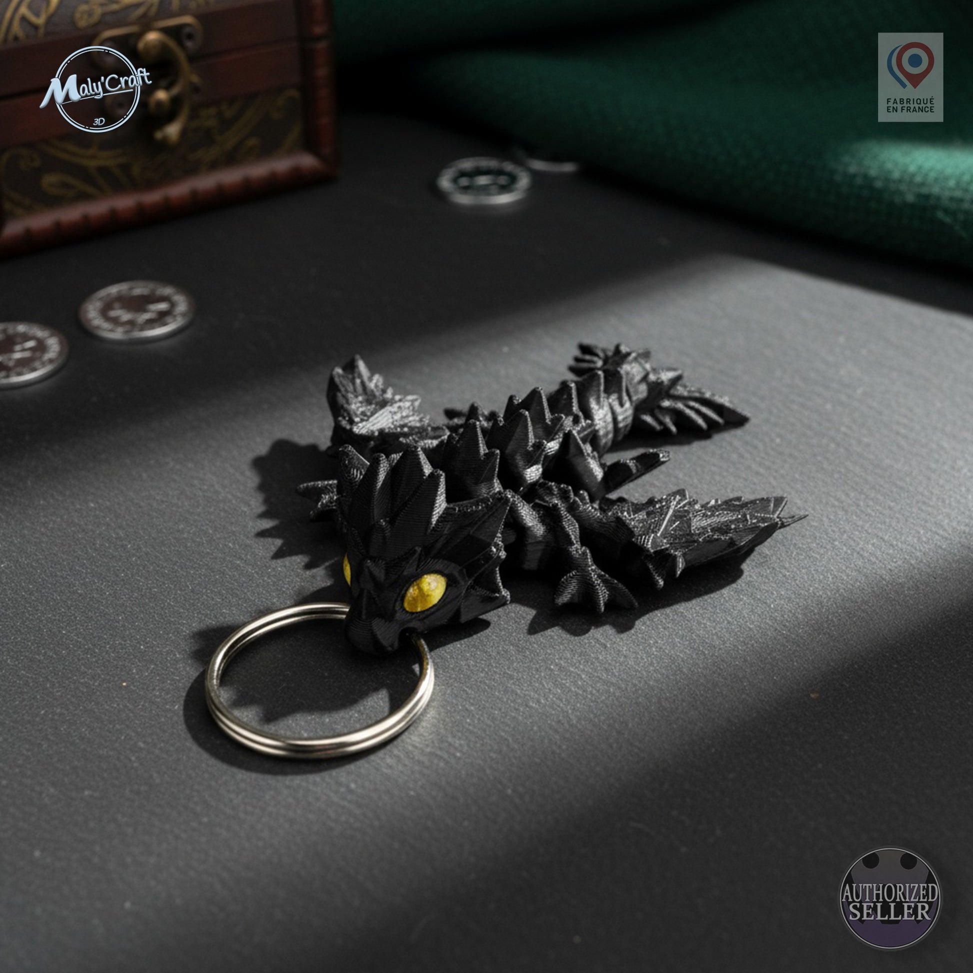Porte-clés dragon articulé imprimé en 3D, de couleur noire, tenu dans la paume d'une main pour montrer sa taille compacte. Le dragon présente des détails texturés avec des yeux jaunes brillants et un aspect écailleux. L’anneau métallique est fixé à la tête de la créature. En arrière-plan, un décor sombre et texturé, avec le logo "MalyCraft" en haut à gauche. Objet original vendu sur Etsy par MalyCraft.
