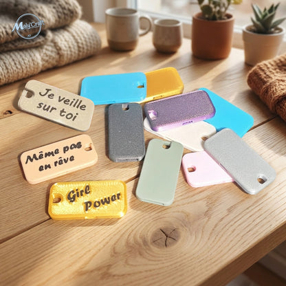 Assortiment de sifflets plats imprimés en 3D de différentes couleurs (bleu, jaune, blanc, gris, violet, vert, rose) posés sur une table en bois, certains personnalisés avec les textes 'Je veille sur toi', 'Même pas en rêve' et 'Girl Power