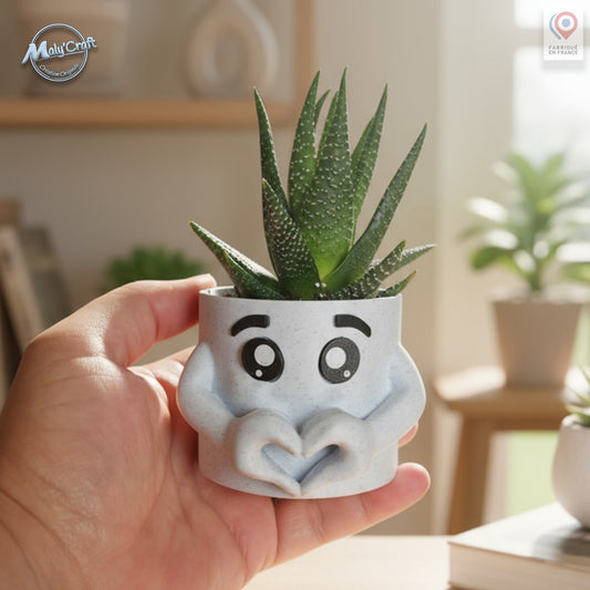 Petit cache-pot en forme de personnage souriant tenant un cœur avec ses mains, contenant une plante succulente. Impression 3D en PLA finition pierre claire. Création originale Maly’Craft, photo prise en lumière naturelle sur un fond d’intérieur doux et lumineux.