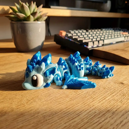 Figurine articulée de dragon de glace imprimée en 3D par Maly'Craft, couleur bleu cristal scintillant. Le dragon est posé sur un bureau en bois clair à côté d'un clavier d'ordinateur noir. Plan moyen horizontal. Fabriqué en France.
