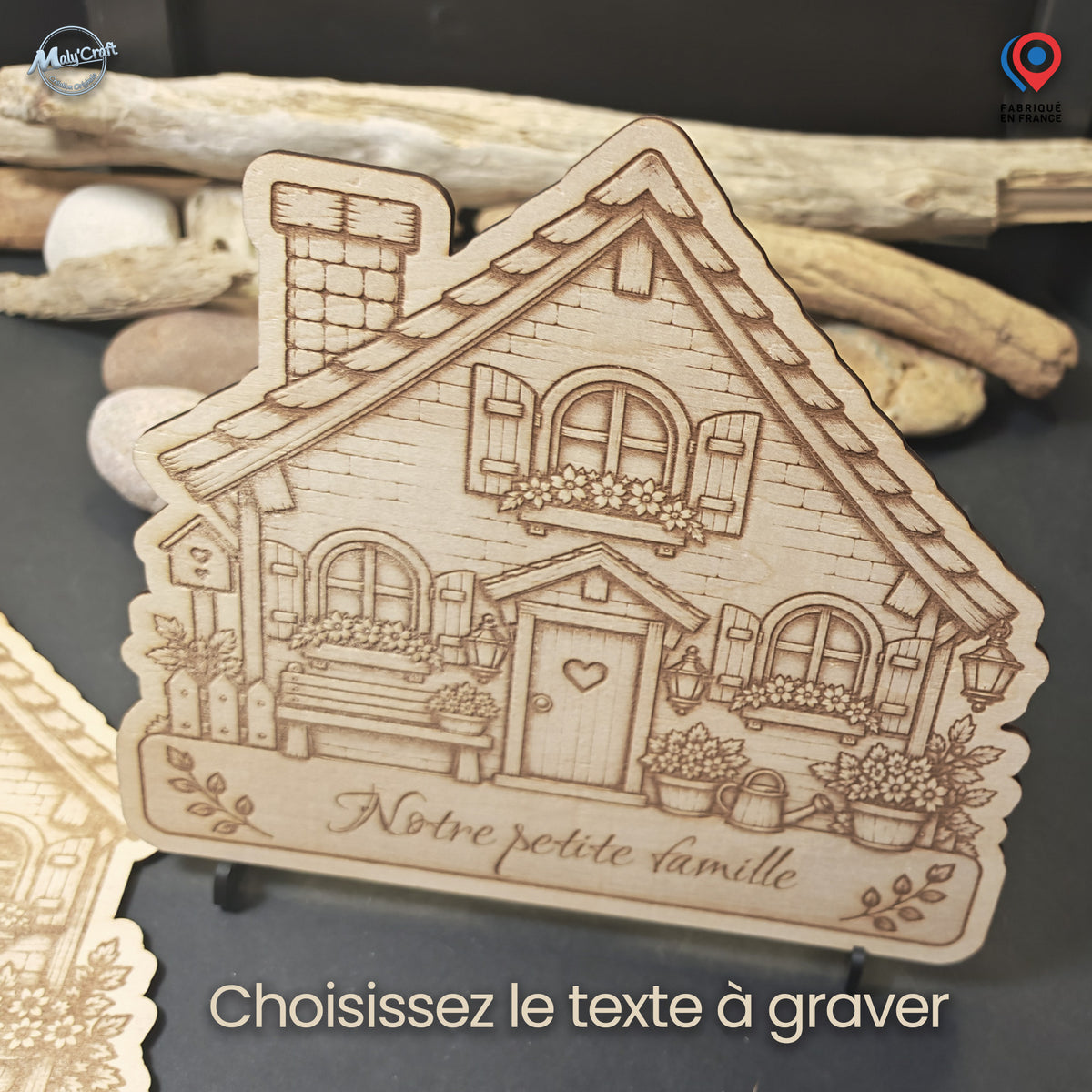 Maison de famille en bois clair montrée sans personnages, sur un support, devant du bois flotté et des galets. La façade gravée présente des fenêtres fleuries, un banc, une porte avec un cœur et un arrosoir. L’inscription Notre petite famille illustre le choix du texte personnalisé à graver.
Light-wood family house plaque shown without portrait figures, standing on a support in front of driftwood and pebbles. The engraved facade features flowered windows, a bench, a heart on the door and a watering can. The