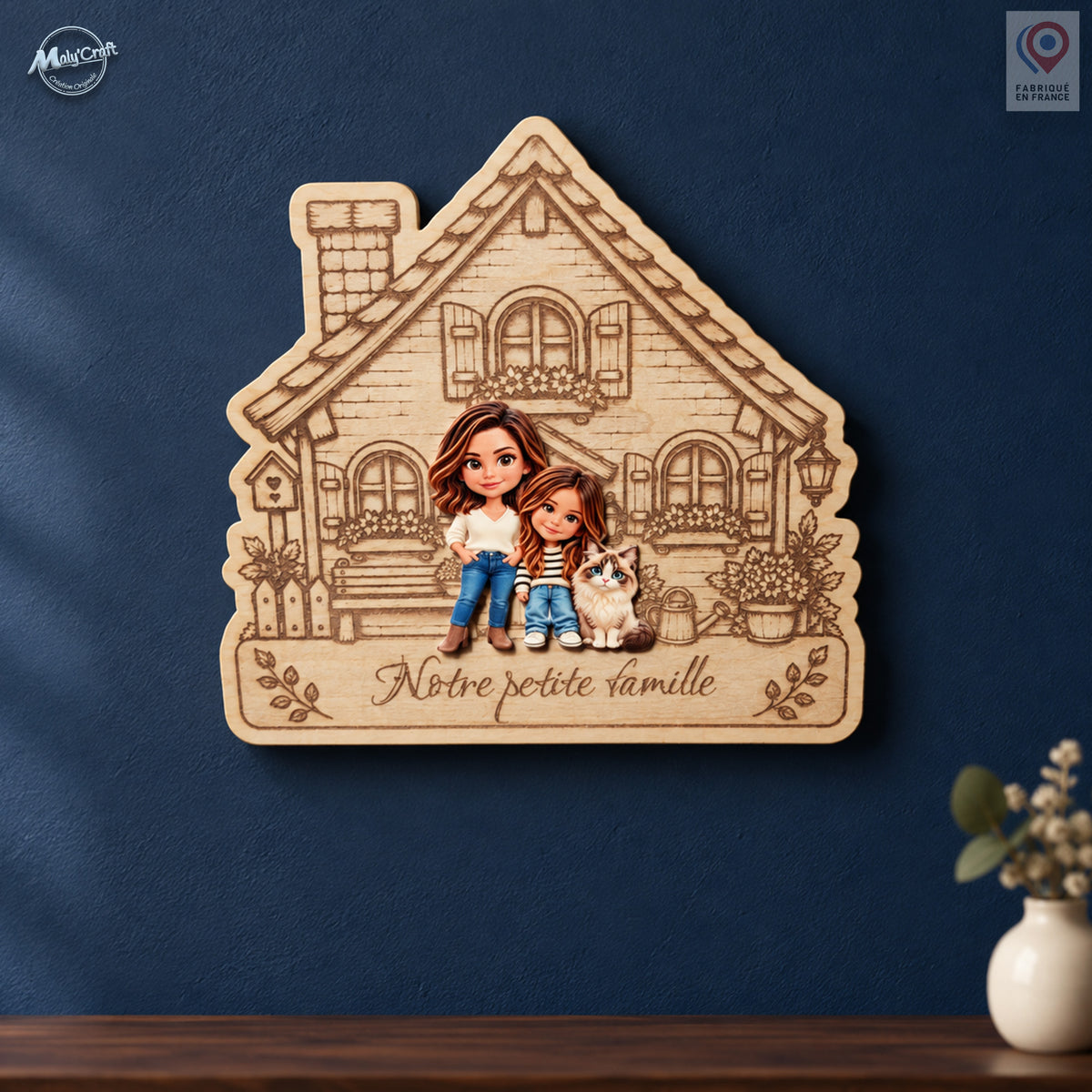 Maison de famille personnalisée en bois clair gravé, ornée des portraits chibi d’une mère, de sa fille et de leur chat. L’inscription Notre petite famille apparaît sous les personnages. Cette décoration murale est présentée sur un mur bleu nuit, au-dessus d’une console en bois avec un vase écru.
Personalized wooden family house plaque featuring colorful chibi portraits of a mother, her daughter and their cat. The French inscription Notre petite famille is engraved beneath the figures. The plaque hangs on a 
