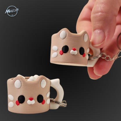 Porte-clés original en forme de petite tasse de café stylisée en tête de chat beige. L'objet présente des détails soignés : de petites oreilles pointues, des joues blanches, des yeux noirs et un petit nez rouge. Une main tient le porte-clés pour montrer sa taille compacte sur un fond noir élégant. Il s'agit d'une impression 3D de précision réalisée avec une finition mate.