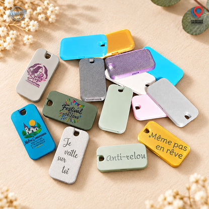 Collection de sifflets d'alerte anti-agression personnalisables MalyCraft, présentés sur un fond beige clair avec une décoration florale discrète. Cet assortiment met en valeur les nombreuses couleurs et finitions disponibles : bleu turquoise, jaune, rose, vert pastel, violet pailleté, blanc nacré et gris métallisé. Plusieurs sifflets présentent des exemples de personnalisation avec des logos d'associations ou de collectivités, des illustrations et des messages tels que « anti-relou », « Je veille sur toi »